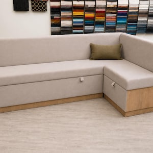 May include: A light beige corner sofa with a wooden base and a matching back. The sofa has a built-in storage compartment with a pull tab. An olive green rectangular cushion rests on the corner. Fabric swatches are in the background.
