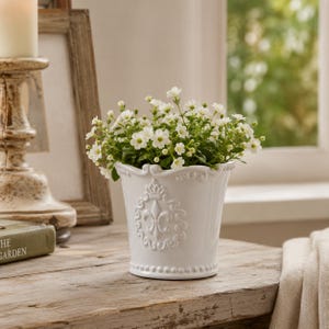 May include: A white ceramic flower pot with a fleur-de-lis design, filled with small white flowers. The pot has a decorative rim and a beaded base. The pot is on a wooden surface.