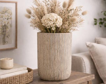 May include: A tall, textured, cylindrical vase filled with dried flowers and grasses. The arrangement includes beige pampas grass, fluffy bunny tails, and a large white hydrangea. The vase sits on a wooden table.