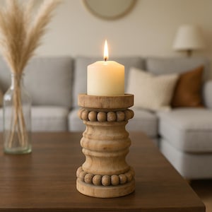 May include: A burning ivory candle on a carved wooden candle holder. The holder features a beaded design and a natural wood finish. The candle sits on a dark brown table, with a blurred living room in the background.