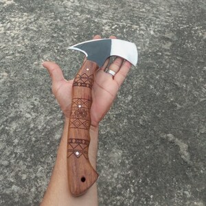 May include: A handcrafted hatchet with a wooden handle featuring intricate geometric carvings. The hatchet head is a combination of black and silver metal. The handle is a warm brown color, and the overall design suggests a rustic, outdoorsy theme.