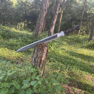 May include: A large, silver sword with a serrated blade and a dark wooden handle rests on a tree stump. The sword's blade has a unique, textured design. The background features a forest with green foliage and tall trees.