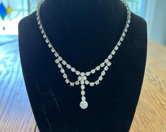 May include: A sparkling silver-toned necklace with clear, round rhinestones. The necklace features a double-strand design at the top, connected by a floral-shaped centerpiece, and a single, dangling rhinestone pendant. The necklace is displayed on a black velvet bust.