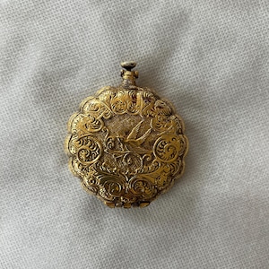 May include: A vintage gold-toned pocket watch with intricate floral and scrollwork. The watch face features a detailed bird design. The watch has a small crown at the top and a hinged opening. The watch is likely made of metal.