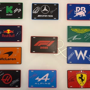 May include: A collection of rectangular license plates featuring Formula 1 team logos and signatures. The plates have various color schemes, including black, blue, orange, and red, with white and colored text and graphics. The plates are arranged on a white surface.