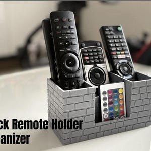 May include: A grey brick-effect remote control tidy holding several black remotes. The organiser has multiple compartments and includes a small remote with coloured buttons. The text "Brick Remote Holder Organizer" is displayed.