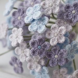 Peut inclure: Gros plan sur un bouquet de fleurs crochetées à la main dans différentes nuances de bleu, de violet et de blanc. Les fleurs ont cinq pétales et un petit centre. Les tiges sont vertes, et l'effet général est délicat et charmant.