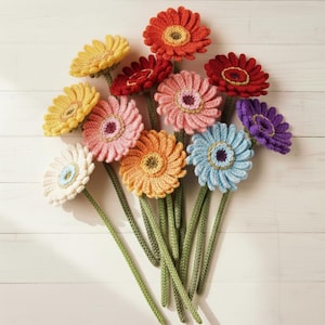 Peut inclure: Un bouquet de gerberas crochetées à la main, aux couleurs variées : jaune, rouge, rose, orange, bleu et violet. Chaque fleur a un centre détaillé et des tiges vertes, disposées sur une surface en bois blanc.