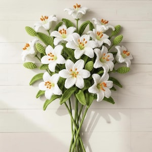 Peut inclure: Un bouquet fait à la main de lys blancs au crochet avec des tiges et des feuilles vertes. Les fleurs ont des centres orange et jaunes. Le bouquet est disposé sur un fond en bois blanc.