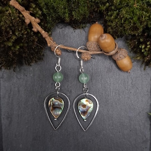 May include: Dangle earrings featuring silver hooks, green beads, and teardrop-shaped iridescent abalone shell pendants. The earrings are presented on a dark grey surface, with acorns and greenery in the background.