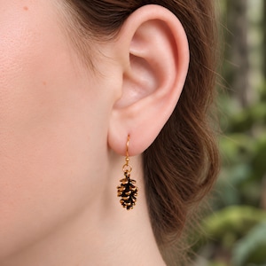 May include: A gold-toned metal earring featuring a small, detailed pinecone charm suspended from a simple hook wire. The pinecone has a textured, layered surface with dark brown accents in the crevices, creating a realistic, three-dimensional appearance.