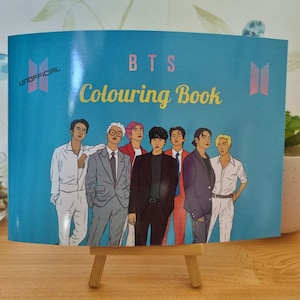 Könnte beinhalten: Ein blaues BTS-Malbuch mit dem Text "BTS Colouring Book" und Illustrationen von sieben Männern. Das Buch wird auf einer kleinen Holzstaffelei präsentiert. Das Wort "UNOFFICIAL" ist links oben aufgedruckt.