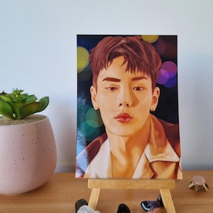 May include: A portrait of a person with auburn hair and a neutral expression, displayed on a small wooden easel. The portrait has a dark background with colorful bokeh effects. The portrait is on a wooden table with decorative stones and a small plant.