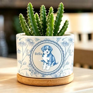 Op de afbeelding: Een witte keramische plantenpot met een blauw bloemenpatroon en een centrale ronde afbeelding van een puppy met de naam Bella. De pot staat op een ronde houten voet en bevat een groene vetplant met stekelige, rechtopstaande stengels.