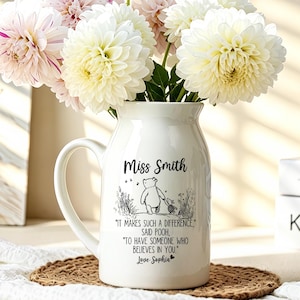 May include: A white ceramic pitcher vase featuring a black ink illustration of Winnie the Pooh and Piglet with the text: Miss Smith, It makes such a difference, said Pooh, to have someone who believes in you, Love Sophia. The vase holds a bouquet of dahlias.