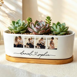 May include: An oval white ceramic planter on a bamboo base containing three green succulents. The front of the planter features a horizontal strip of four printed photographs of a couple, with the names "daniel" and "sophia" written in black script.