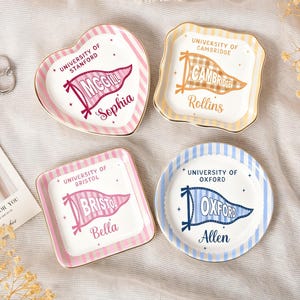 Può includere: Quattro piatti decorativi in ceramica di varie forme: cuore, quadrato e rotondo. Ogni piatto presenta il nome di un'università, un design a gagliardetto e un nome. I piatti hanno bordi dorati e motivi a righe rosa, gialle e blu.
