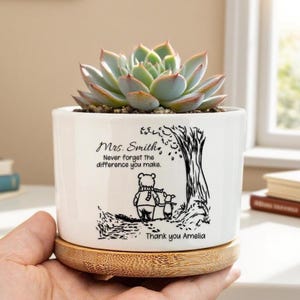Peut inclure: Un pot de fleurs en céramique blanche avec une plante succulente. Le pot présente une illustration noire d'un ours et d'un porcelet sous un arbre, avec le texte "Mrs. Smith, N'oubliez jamais la différence que vous faites." Un dessous de plat en bambou soutient le pot.