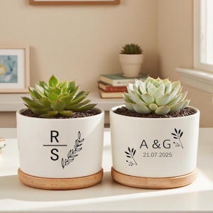 May include: Two white ceramic plant pots with succulents, each on a wooden base. One pot has the initials "R S" and a leaf design. The other has "A & G" and the date "21.07.2025".