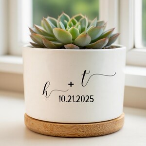 Pode incluir: Um vaso de cerâmica branco com uma planta suculenta. O vaso tem as letras "h + t" e a data "10.21.2025" em escrita preta. Assenta numa base redonda de madeira natural. A suculenta tem folhas verdes e verde claro.