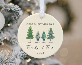 May include: A round, cream-colored Christmas ornament with a white ribbon. The ornament features four illustrated Christmas trees and the text "FIRST CHRISTMAS AS A Family of Four -2024-". The names "DAD", "EMMA", "MAX", and "MOM" are below the trees.