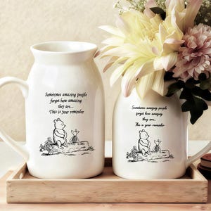 May include: Two white ceramic vases with handles, each decorated with a Winnie the Pooh illustration and the text "Sometimes amazing people forget how amazing they are... This is your reminder." One vase contains flowers. They sit on a wooden tray.