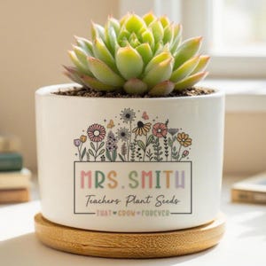 Może przedstawiać: Biała ceramiczna doniczka z rośliną sukulentową. Doniczka ma kwiatowy wzór i napis "MRS. SMITH Teachers Plant Seeds". Doniczka stoi na okrągłym drewnianym spodku.