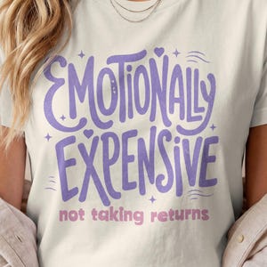 Puede incluir: Una camiseta de manga corta de color crema con el texto morado "EMOTIONALLY EXPENSIVE" en una fuente manuscrita estilizada, acompañada de pequeñas estrellas y corazones, con el texto rosa "not taking returns" impreso debajo.