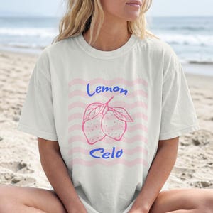 May include: White t-shirt with a pink and blue lemon graphic. The design features two lemons with leaves, the word "Lemon" above, and "Celo" below, set against a pink wavy background. The shirt is worn on a beach.