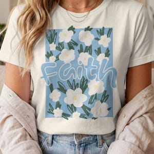 Puede incluir: Camiseta color crema con un diseño floral. Flores blancas y hojas verdes sobre un fondo azul claro. La palabra "Faith" está escrita en cursiva azul claro sobre el diseño.