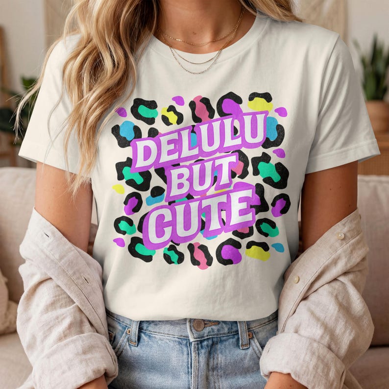 Puede incluir: Camiseta color crema con el texto "DELULU BUT CUTE" en fuente morada y blanca. La camiseta presenta un estampado de leopardo colorido en negro, verde, azul, amarillo y rosa.
