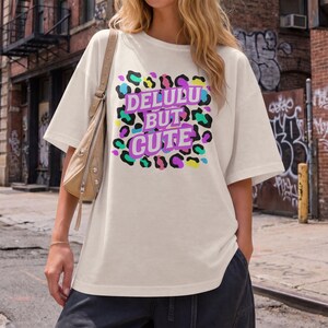 Puede incluir: Camiseta color crema con el texto "DELULU BUT CUTE" en un dise&ntilde;o de estampado de leopardo colorido. La camiseta tiene mangas cortas y un corte holgado, adecuada para uso casual.