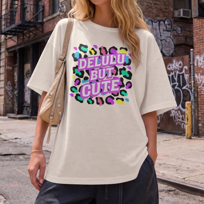 Puede incluir: Camiseta color crema con el texto "DELULU BUT CUTE" en un dise&ntilde;o de estampado de leopardo colorido. La camiseta tiene mangas cortas y un corte holgado, adecuada para uso casual.