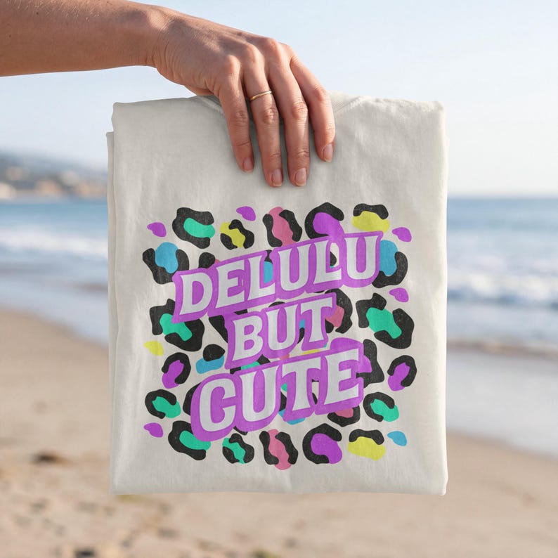 Puede incluir: Camiseta color crema con el texto "DELULU BUT CUTE" en morado, negro y blanco. El texto est&aacute; rodeado de un estampado de leopardo en turquesa, azul, amarillo y morado.