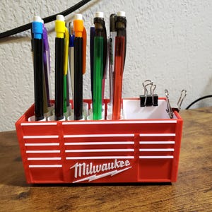 May include: Red tool box-style pen holder with the word "Milwaukee" in white letters. The holder has 12 slots for pens and pencils, and is filled with a variety of colored pens and pencils.