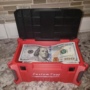 May include: A red storage box with a black lid, open to reveal several &pound;100 bills. The box has the words "Custom Text" printed on the front. The box is sitting on a speckled surface.