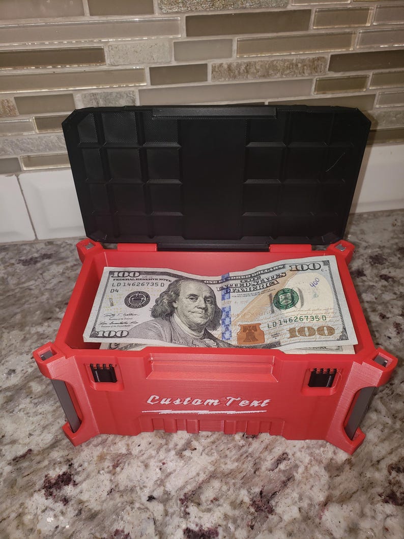 May include: A red storage box with a black lid, open to reveal several &pound;100 bills. The box has the words "Custom Text" printed on the front. The box is sitting on a speckled surface.