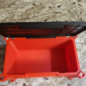 May include: An open, orange plastic toolbox with a black lid. The interior of the toolbox is visible, showing its empty space. The toolbox has a rectangular shape with reinforced corners and a textured surface.