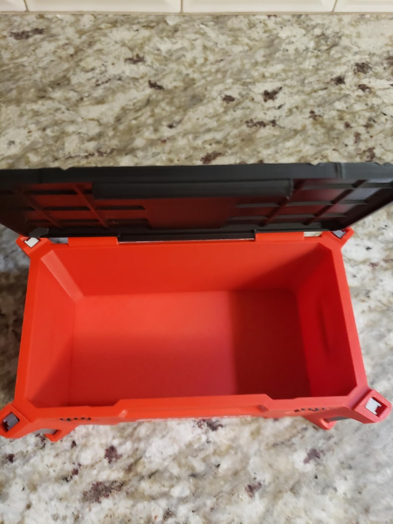 May include: An open, orange plastic toolbox with a black lid. The interior of the toolbox is visible, showing its empty space. The toolbox has a rectangular shape with reinforced corners and a textured surface.