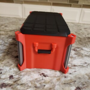 May include: A red and black rectangular tool box with a black handle and a textured black lid. The tool box has a red body with a handle. It is on a speckled surface.