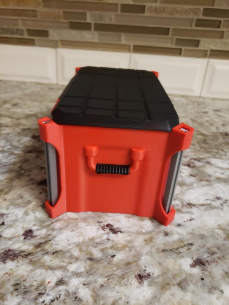 May include: A red and black rectangular tool box with a black handle and a textured black lid. The tool box has a red body with a handle. It is on a speckled surface.