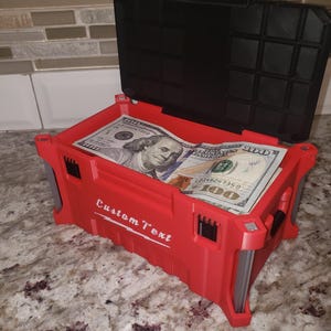 May include: A red storage box with a black lid, filled with U.S. $100 bills. The box has the words "Custom Text" printed on the side. The box is open, revealing the money inside. It sits on a speckled countertop.