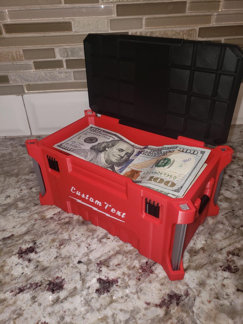 May include: A red storage box with a black lid, filled with U.S. $100 bills. The box has the words "Custom Text" printed on the side. The box is open, revealing the money inside. It sits on a speckled countertop.