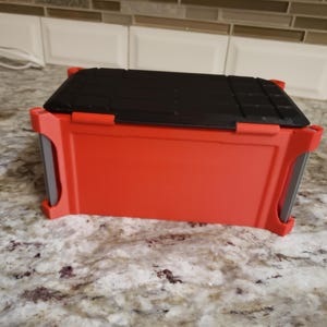 May include: A red plastic toolbox with a black lid. The toolbox is rectangular with rounded corners and a handle on each side. The lid is textured with parallel lines. The toolbox sits on a granite countertop. The dimensions are approximately 25 cm x 15 cm x 15 cm.