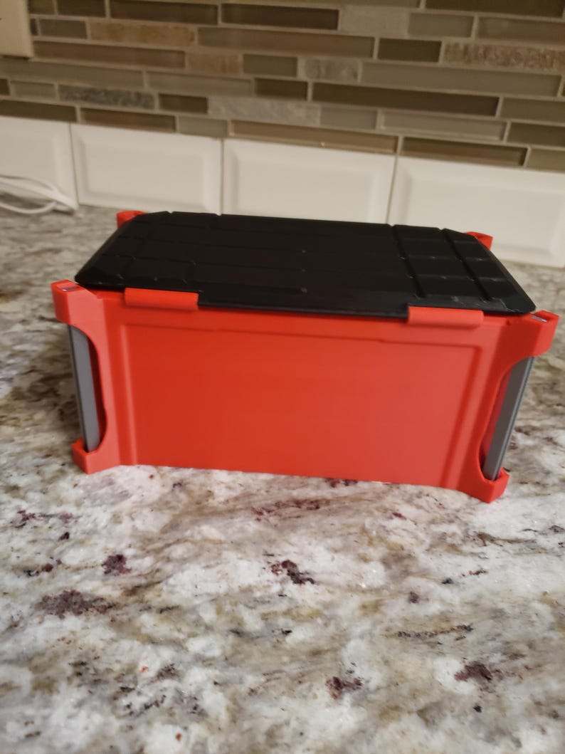 May include: A red plastic toolbox with a black lid. The toolbox is rectangular with rounded corners and a handle on each side. The lid is textured with parallel lines. The toolbox sits on a granite countertop. The dimensions are approximately 25 cm x 15 cm x 15 cm.