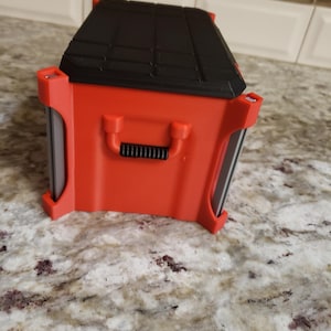 May include: A red and black toolbox with a handle. The toolbox has a black, textured lid and red sides. The handle is black and ribbed. The toolbox is on a speckled countertop.