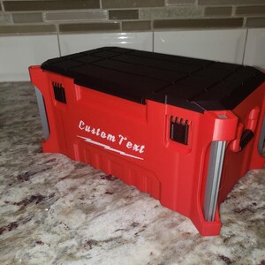 May include: A red toolbox with a black lid and the words "Custom Text" in white. The toolbox is rectangular with a textured surface and metal handles. It sits on a marble countertop. The toolbox is approximately 30 cm long.