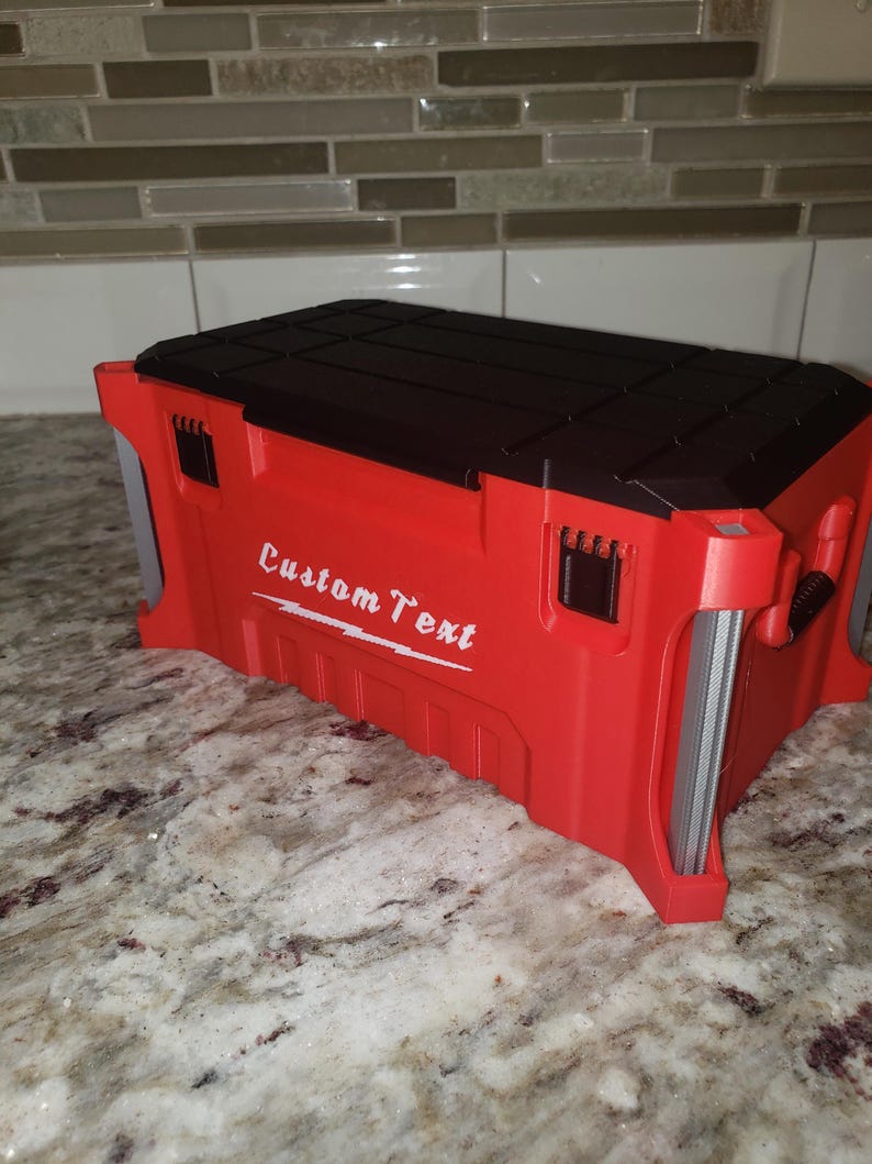 May include: A red toolbox with a black lid and the words "Custom Text" in white. The toolbox is rectangular with a textured surface and metal handles. It sits on a marble countertop. The toolbox is approximately 30 cm long.