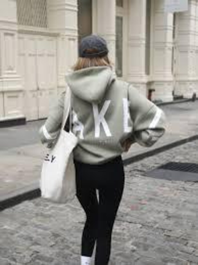 May include: A light green hooded sweatshirt with the word "AKEY" printed in white on the back. The sweatshirt has white stripes on the sleeves and is being worn with black leggings.