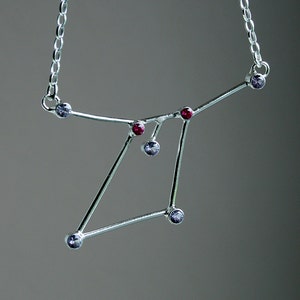 Op de afbeelding: Een zilveren ketting met een sterrenbeeldontwerp met kleine paarse en rode edelstenen.